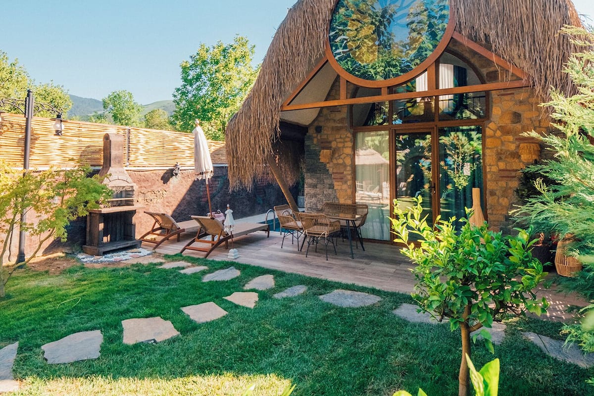 Uma casa com telhado de palha, janela redonda, paredes de pedra e um quintal gramado com móveis de exterior.