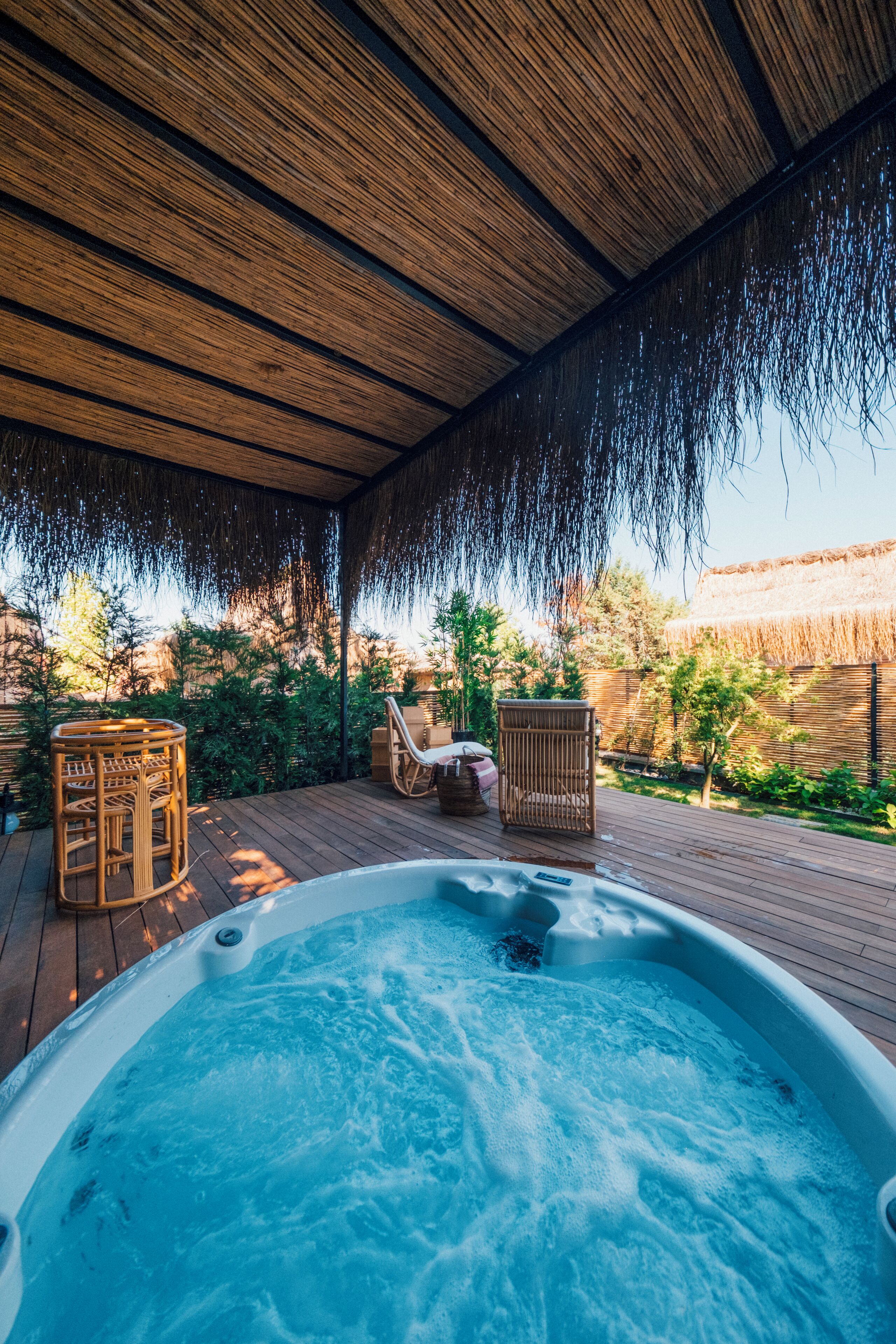 Un bain à remous sur une terrasse en bois, avec des fauteuils en osier et un toit de chaume au-dessus.
