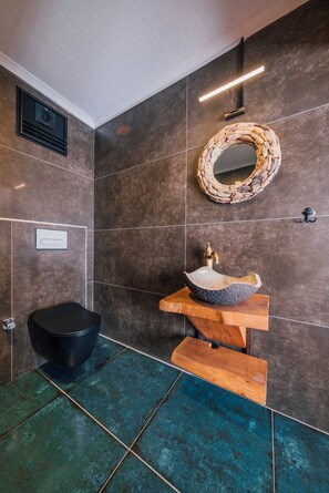 A bathroom with dark tiled walls, a round mirror, a wooden vanity, and a black toilet. - Feronia Suites (Kartepe)