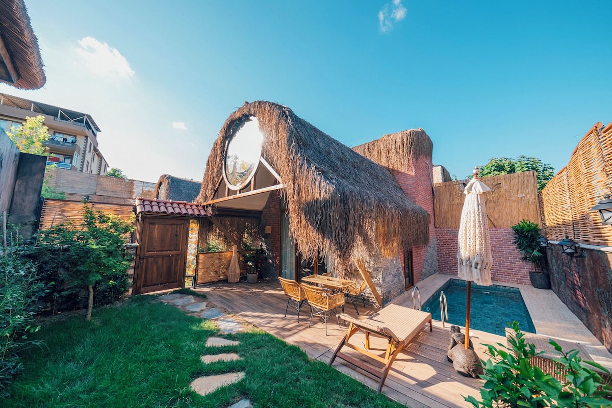 Uma estrutura com telhado de palha, mesa e cadeiras de madeira, piscina e área de jardim com deck e pedras no caminho.