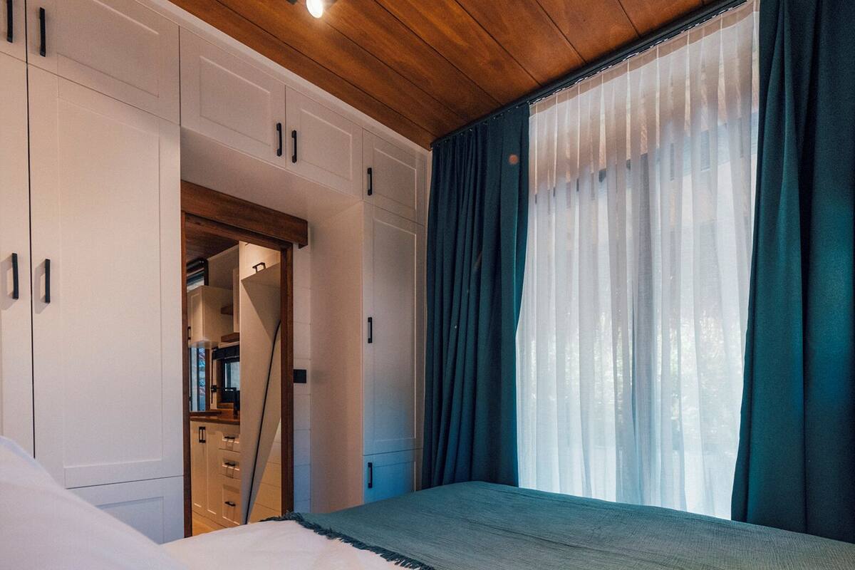 Quarto com teto de madeira, cama com roupa de cama branca, guarda-roupa com portas brancas e janela com cortinas azuis.