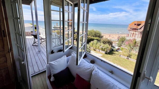 Habitación con vista a la playa y al océano, un asiento junto a la ventana con cojines, y una terraza de madera afuera.