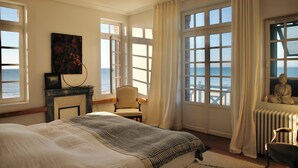 Un dormitorio con una cama grande, una silla, chimenea y vista al mar.