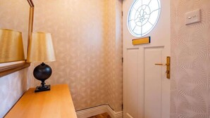 Un pasillo con papel tapiz estampado, una mesa de madera con una lámpara, y una puerta con un ventanal circular.