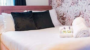 Una cama bien hecha con almohadas blancas y negras, un cabecero tapizado en rosa y un fondo de papel pintado con motivos florales.