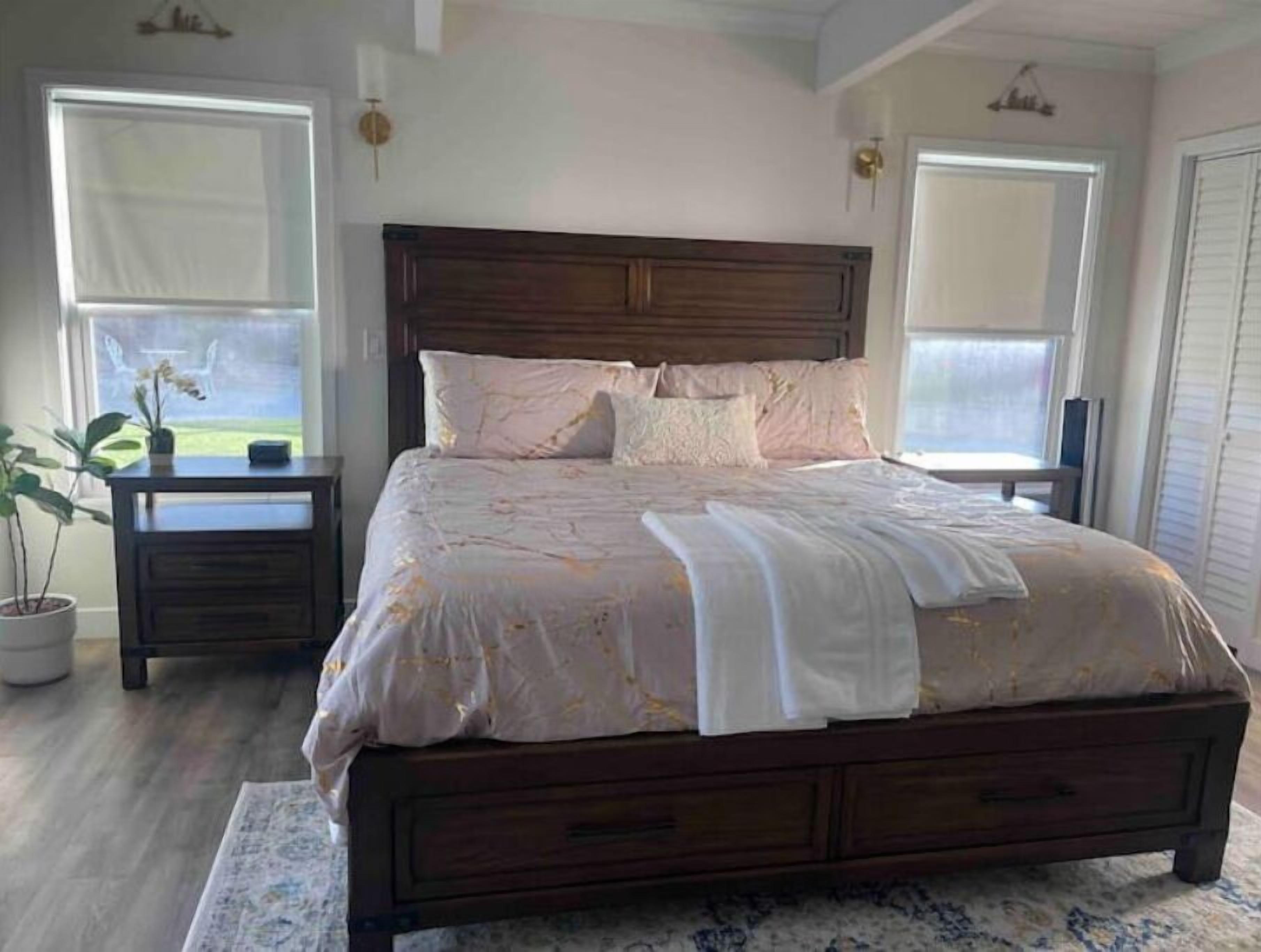 Un dormitorio con una cama de madera, dos mesitas de noche, una planta en maceta y vistas al exterior a través de ventanas amplias.