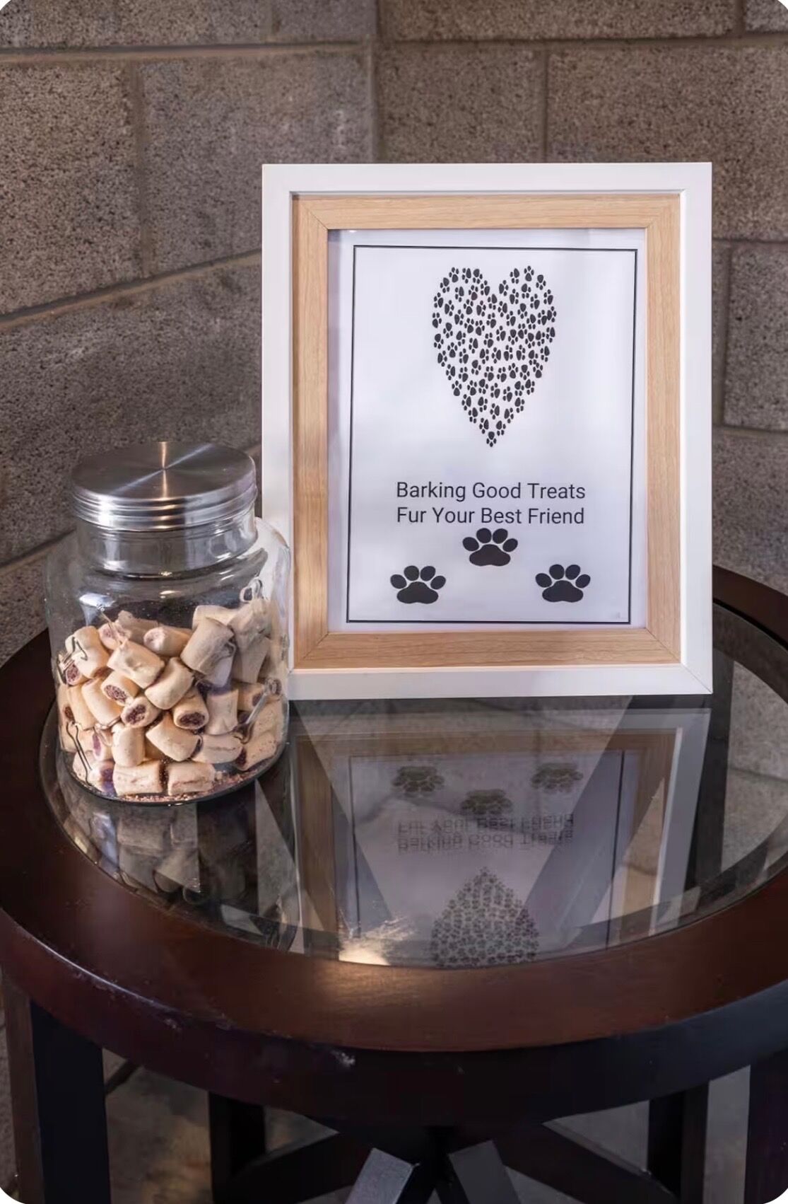 A framed picture with a heart-shaped paw print design and text, a jar of treats, and a glass table.