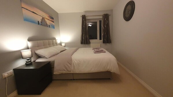 A bedroom with a bed, bedside tables, a nightstand, a painting, and a window with curtains.