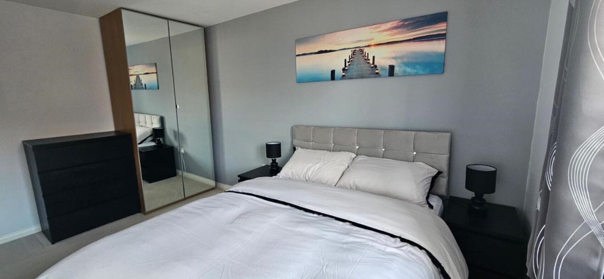 A hotel room with a large bed, a nightstand with a lamp, a mirror, and a painting on the wall.