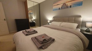 A neatly made bed with folded towels, a large mirror, and a painting of a sunset on the wall.