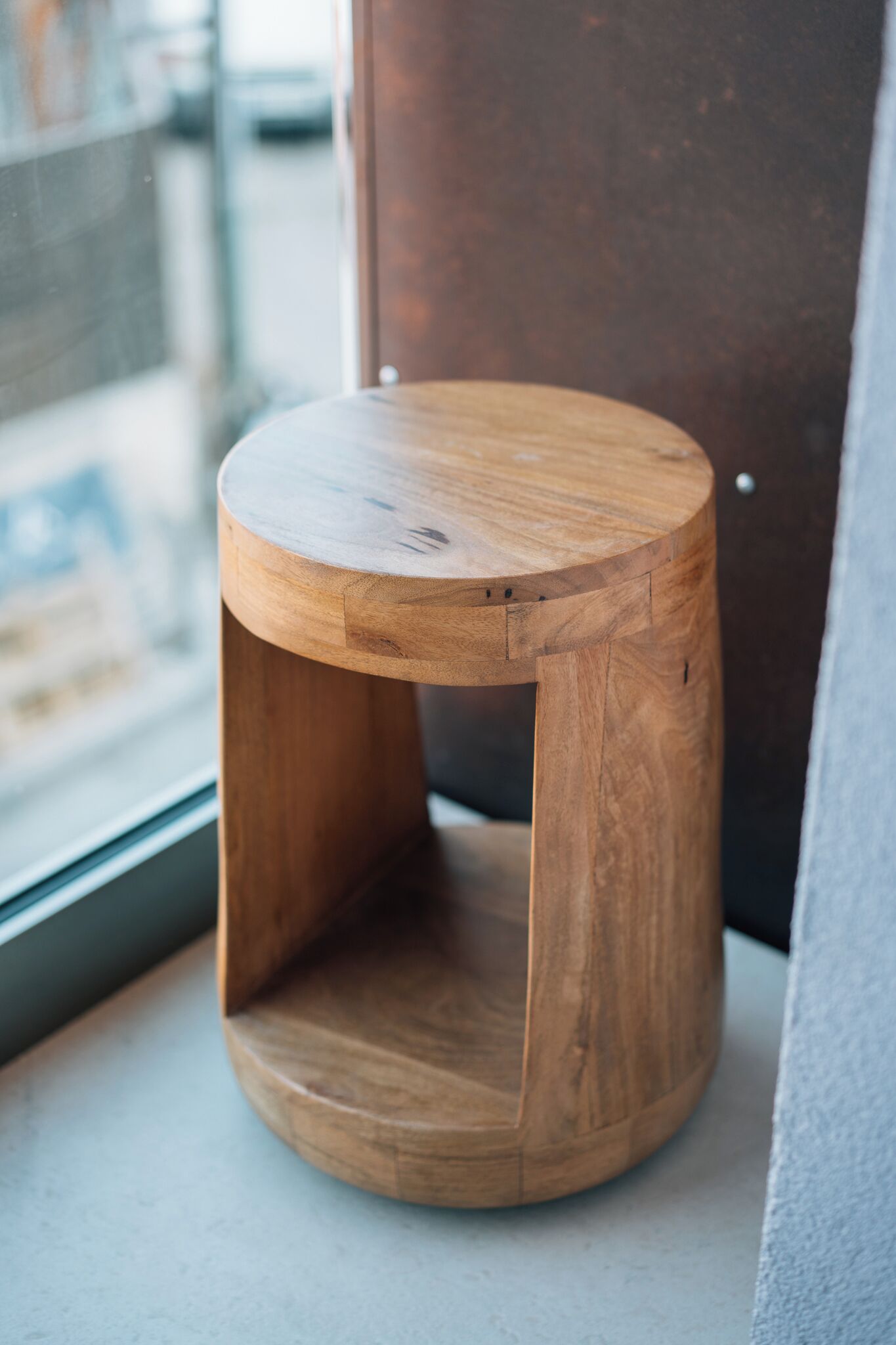 A wooden stool with a round top and a cut-out section, placed on a windowsill.