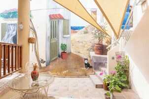 Una mesa de vidrio con un jarrón, un gato y plantas en macetas en un balcón con vista a un paisaje pintado.