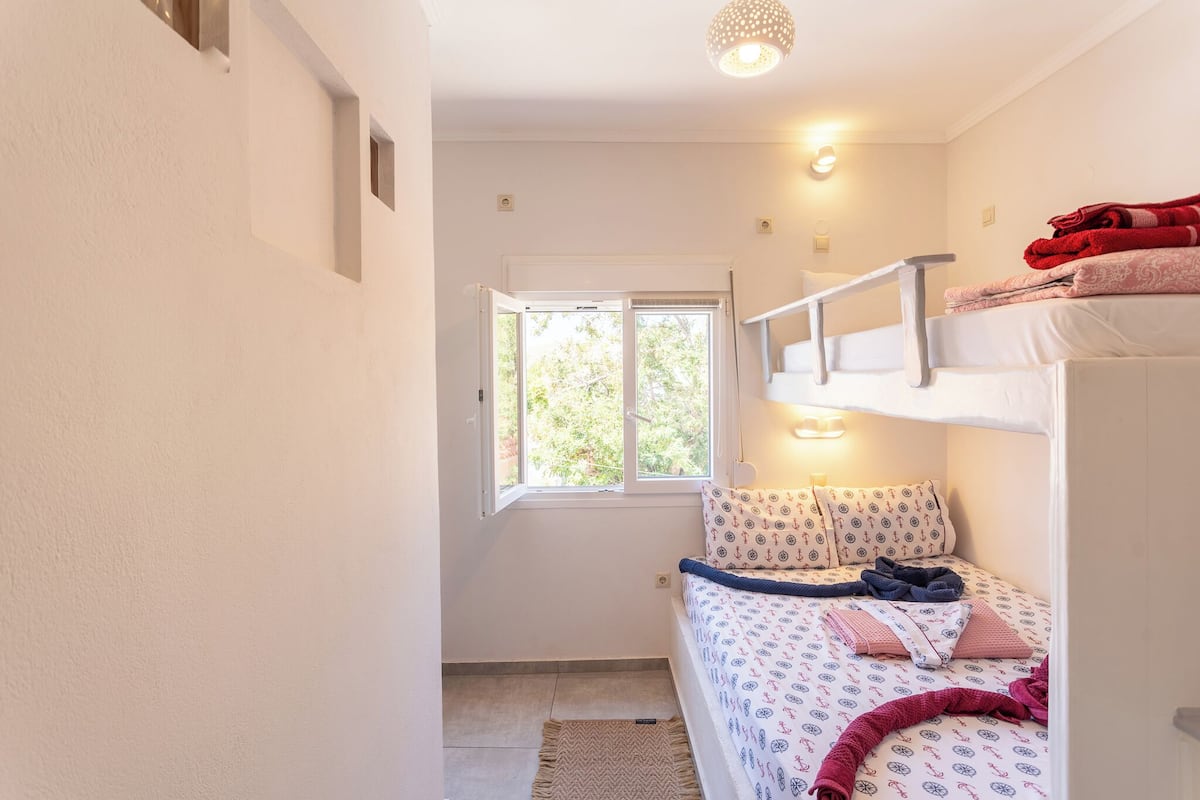 A small, well-lit bedroom with a bunk bed, a window with a view of greenery, and a bedside table with a lamp.