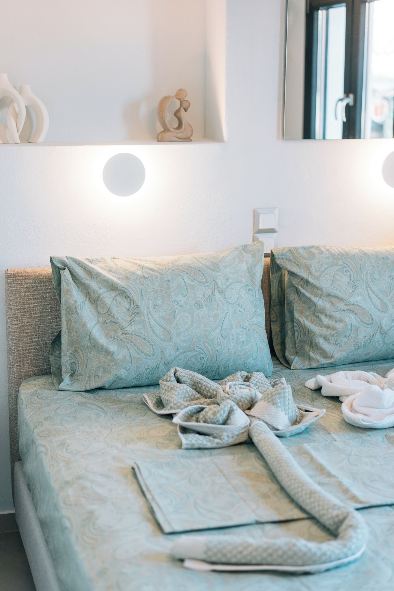A neatly made bed with patterned bedding, a bedside table with a lamp, and a wall-mounted mirror.