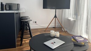 A modern kitchen with a refrigerator, microwave, and coffee maker. There is a black table with a laptop and books, and a large flat-screen TV on a tripod stand.