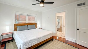 Une chambre à coucher avec un lit, un ventilateur de plafond, une table de chevet, une fenêtre et une porte.