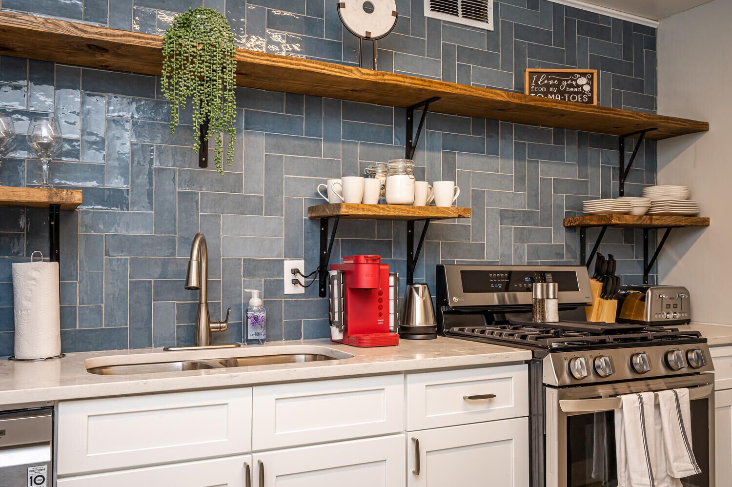 A kitchen with blue tiled backsplash, white cabinets, a stainless steel sink, a red coffee maker, and a gas stove.
