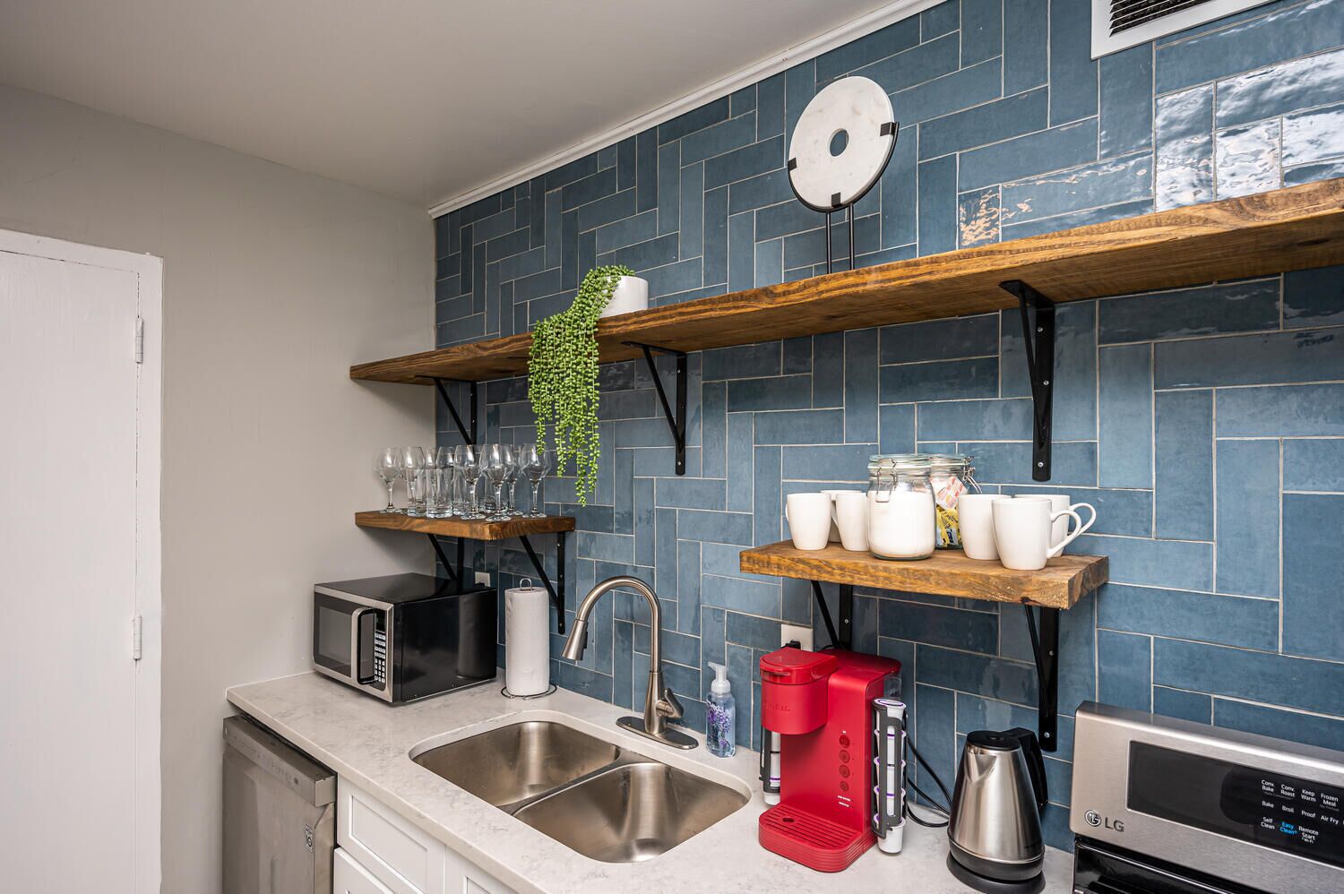 A modern kitchen with a blue tiled backsplash, stainless steel appliances, and wooden shelves holding glassware and kitchen items.