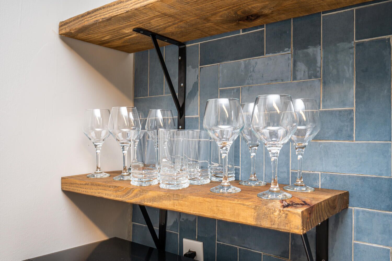 A wooden shelf with multiple wine glasses against a tiled wall.