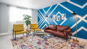 Un salon avec un mur orné d’un motif à chevrons bleus et blancs, un canapé en cuir marron, des fauteuils jaunes, une table basse en verre et un tapis coloré.