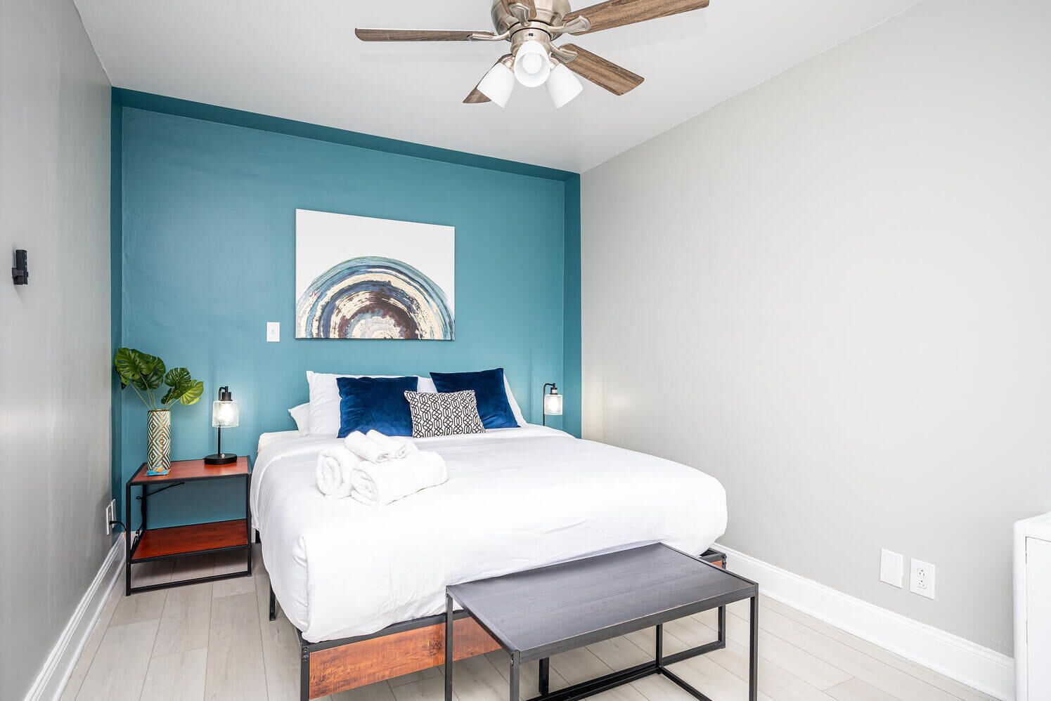 Un dormitorio moderno con una pared de acento color aguamarina, una cama con ropa de cama blanca, una mesita de noche de madera y un ventilador de techo.