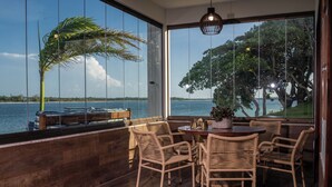 Comedor con sillas de mimbre y una mesa de madera, con vistas a un cuerpo de agua y un árbol.