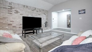 Un salon moderne avec un mur d’accent en briques, un téléviseur à écran plat, une table basse en verre et un canapé avec des coussins.