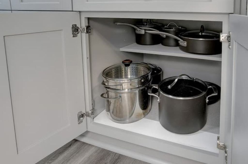 Un meuble de cuisine contenant des ustensiles de cuisine en acier inoxydable, notamment des casseroles et des poêles, ainsi qu’une passoire.