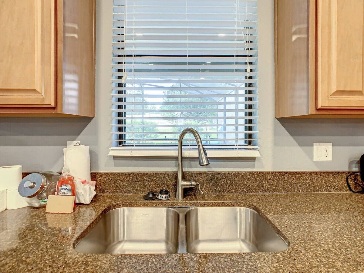 A kitchen with a stainless steel sink, granite countertop, wooden cabinets, and a window with blinds.