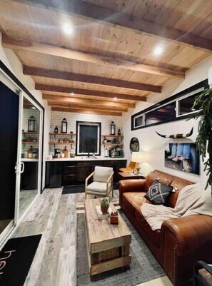 A cozy living room with a brown leather sofa, a wooden coffee table, and a kitchen area with a brick backsplash and wooden beams. - Peaceful new Tiny Home on farm, steps to wineries!
(Escondido)