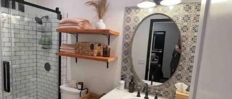 Un baño moderno con cabina de ducha de vidrio, inodoro blanco, tocador con un espejo grande y estantes de madera sobre el lavamanos.