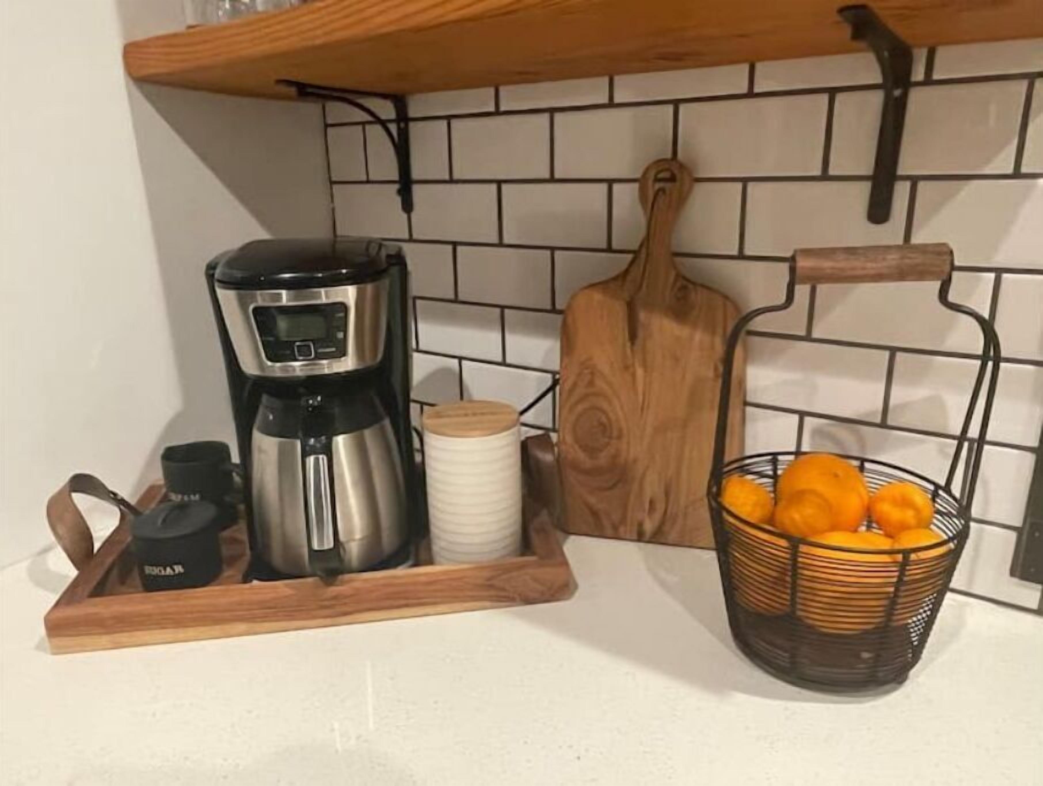 Una encimera de cocina con una cafetera, una tabla de cortar, una cesta de naranjas y una bandeja de madera con un recipiente.