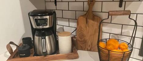 Una encimera de cocina con una cafetera, una tabla de cortar, una cesta de naranjas y una bandeja de madera con un recipiente.