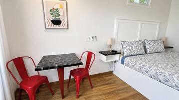 Una habitación pequeña y limpia con una cama, una mesita de noche, una lámpara, un cuadro enmarcado y dos sillas rojas alrededor de una mesa.