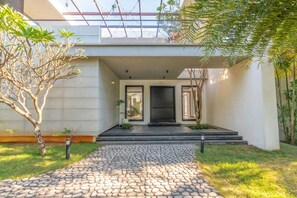 Una casa moderna con un camino de piedra que conduce a una puerta negra, rodeada de follaje y una estructura de techo de cristal.