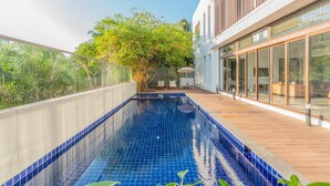 A rooftop swimming pool with blue tiles, wooden deck, and a view of a modern building with large windows. - Truliv Villa LAGUNA (Tirupporur)