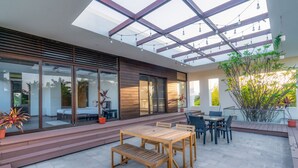 A modern outdoor patio with a wooden table and chairs, a glass door leading to a room, and a pergola with string lights. - Truliv Villa LAGUNA (Tirupporur)