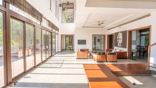 A spacious living area with large windows, a wooden floor, and a modern sofa. - Truliv Villa LAGUNA (Tirupporur)