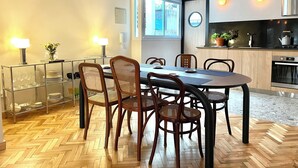 Un espace repas moderne avec un parquet en bois disposé en chevrons, une table ronde et des chaises en osier. On y trouve une rambarde en verre avec un cadre en métal, une plante en pot et une vue sur la cuisine, où l’on aperçoit une cuisinière et un four.