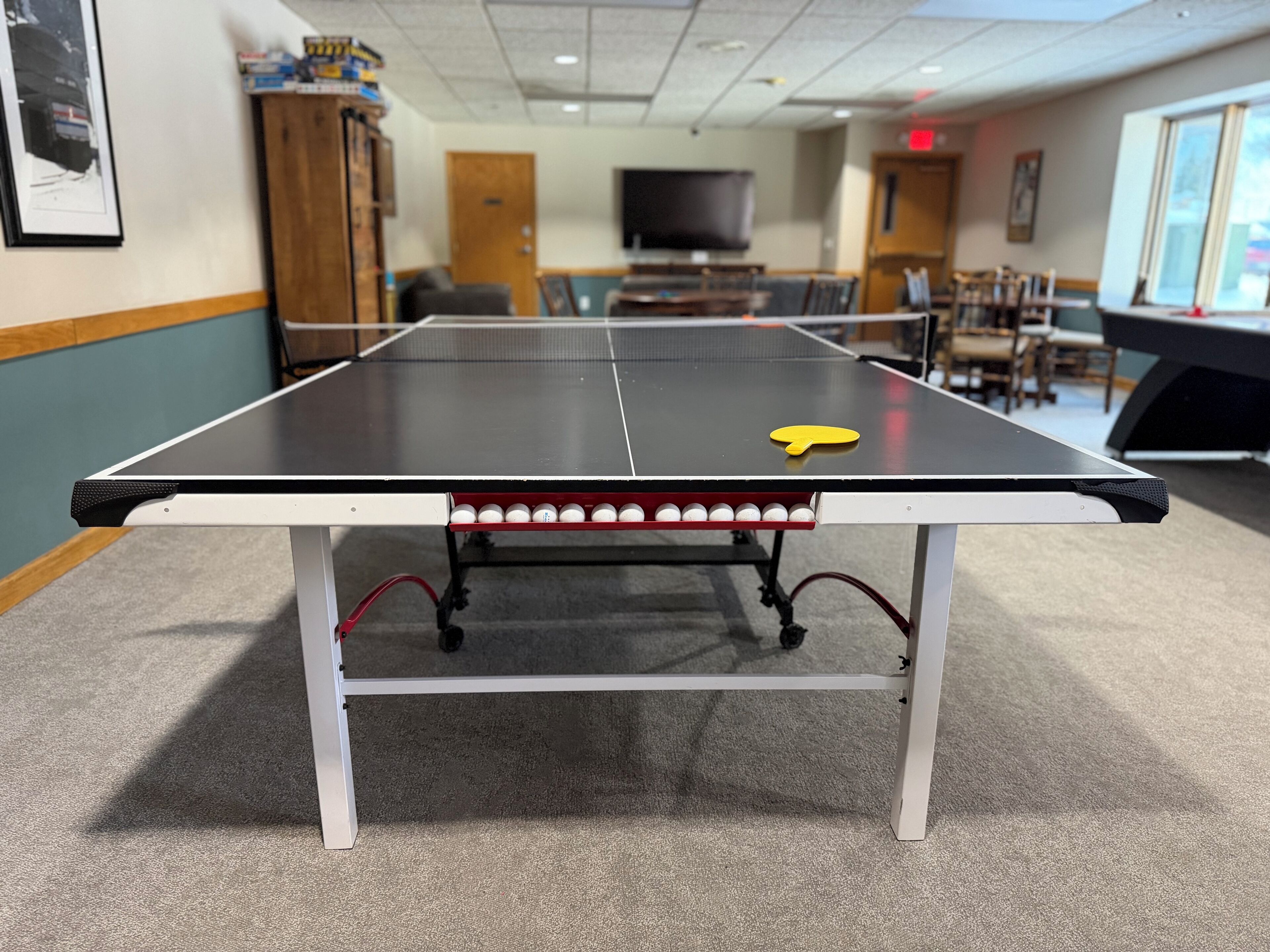 Une table de ping-pong avec une raquette jaune au centre, entourée de chaises et avec un téléviseur en arrière-plan.