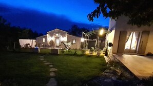 Un'area esterna ben illuminata con un vialetto in pietra che conduce a un edificio, circondata da giardini curati e un cielo sereno.