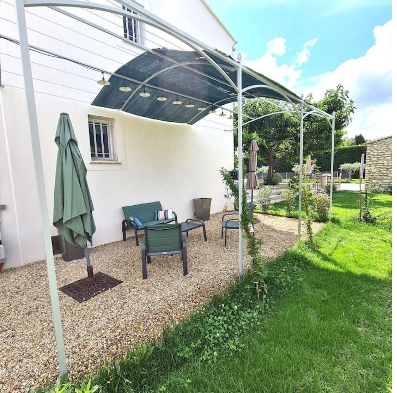 Een overdekt terras met tuinmeubilair, een groene parasol en een tuin.