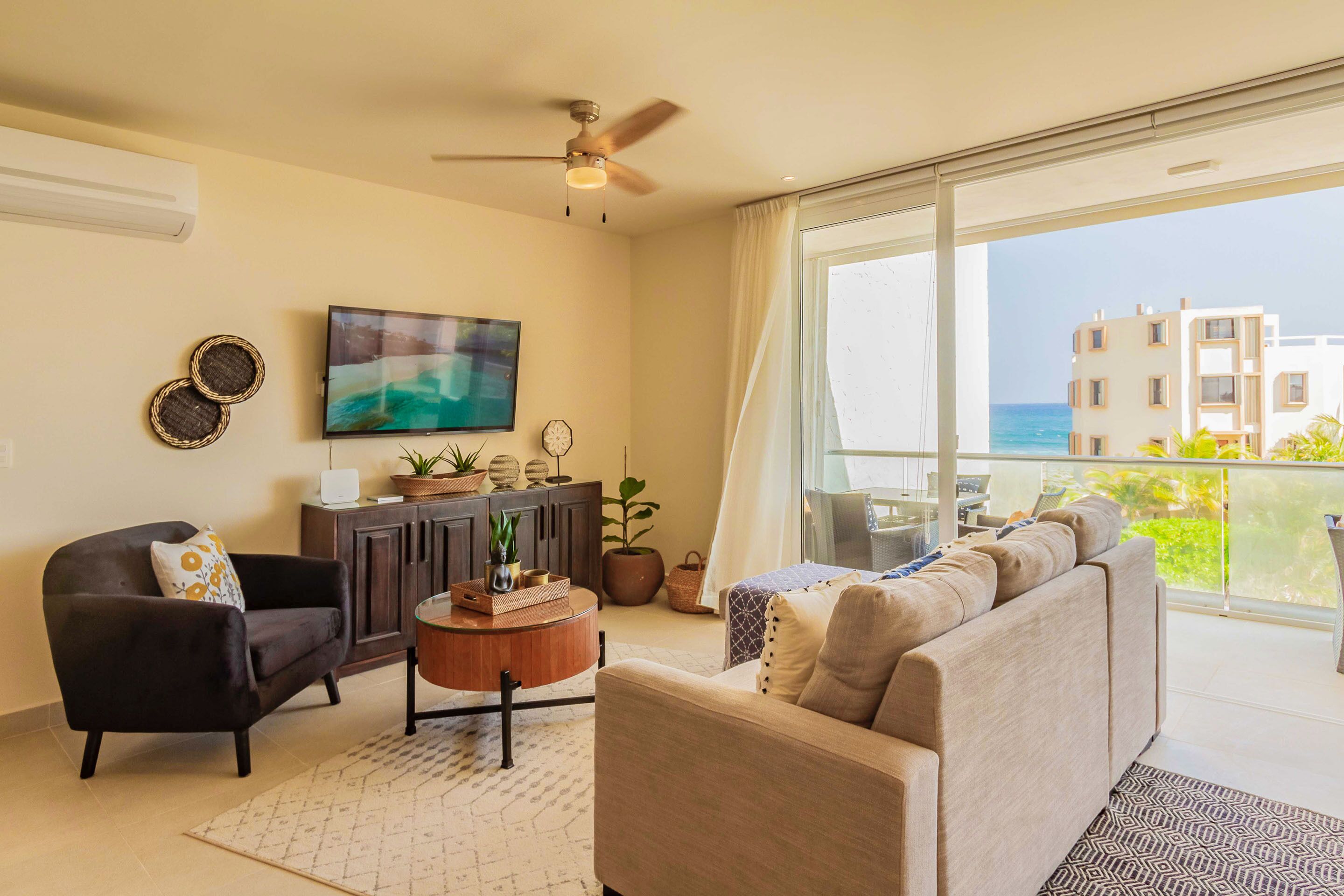 A living room with a sofa, armchair, and coffee table, a TV, and a balcony with a view of buildings and the sea.