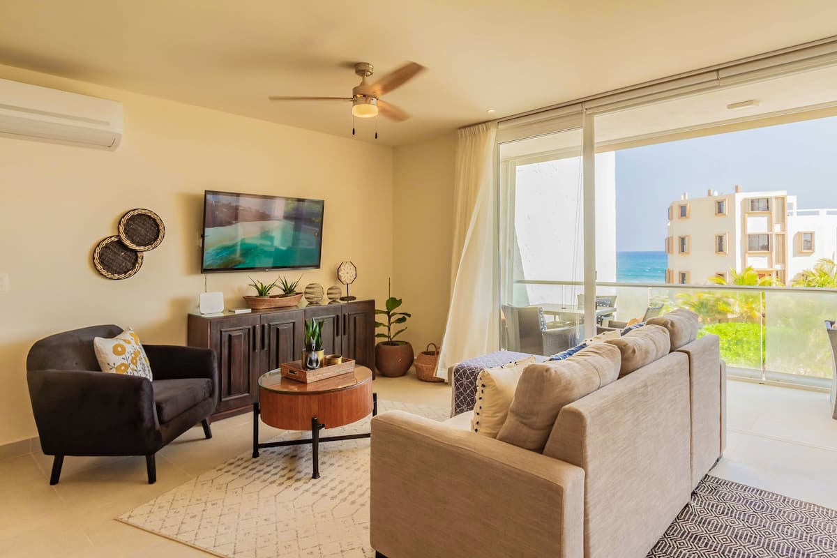 A living room with a sofa, armchair, and coffee table, a TV, and a balcony with a view of buildings and the sea.