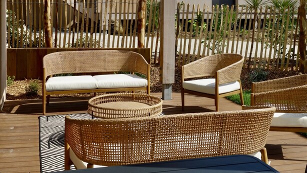 A covered outdoor seating area with wicker furniture, a staircase leading to an upper level, and a building with a dark shuttered window. - Maison Décorée Avec Charme, Parfaitement Équipée Pour 6 Personne, Proche Plage (Barneville-Carteret)