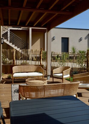 A covered outdoor seating area with wicker furniture, a staircase leading to an upper level, and a building with a dark shuttered window. - Maison Décorée Avec Charme, Parfaitement Équipée Pour 6 Personne, Proche Plage (Barneville-Carteret)