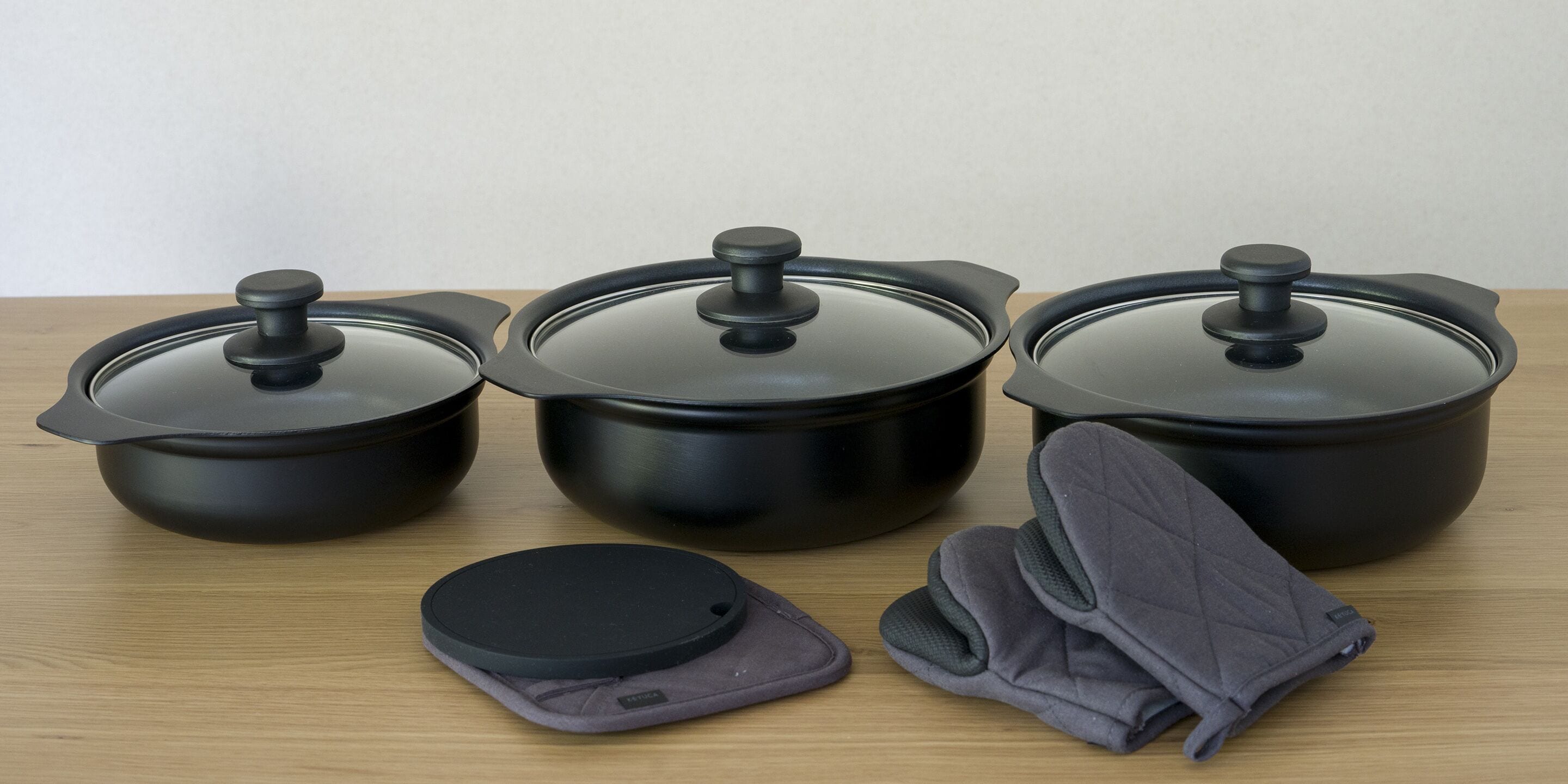 Three black pots with lids and a pair of gray oven mitts on a wooden surface.