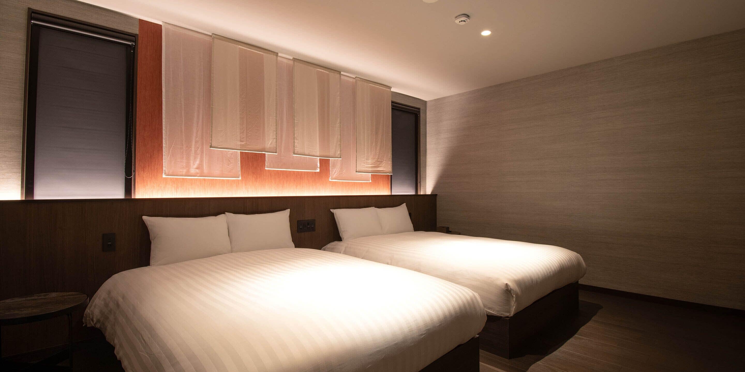 A modern hotel room with two beds, a wooden headboard, and a large window with blinds.