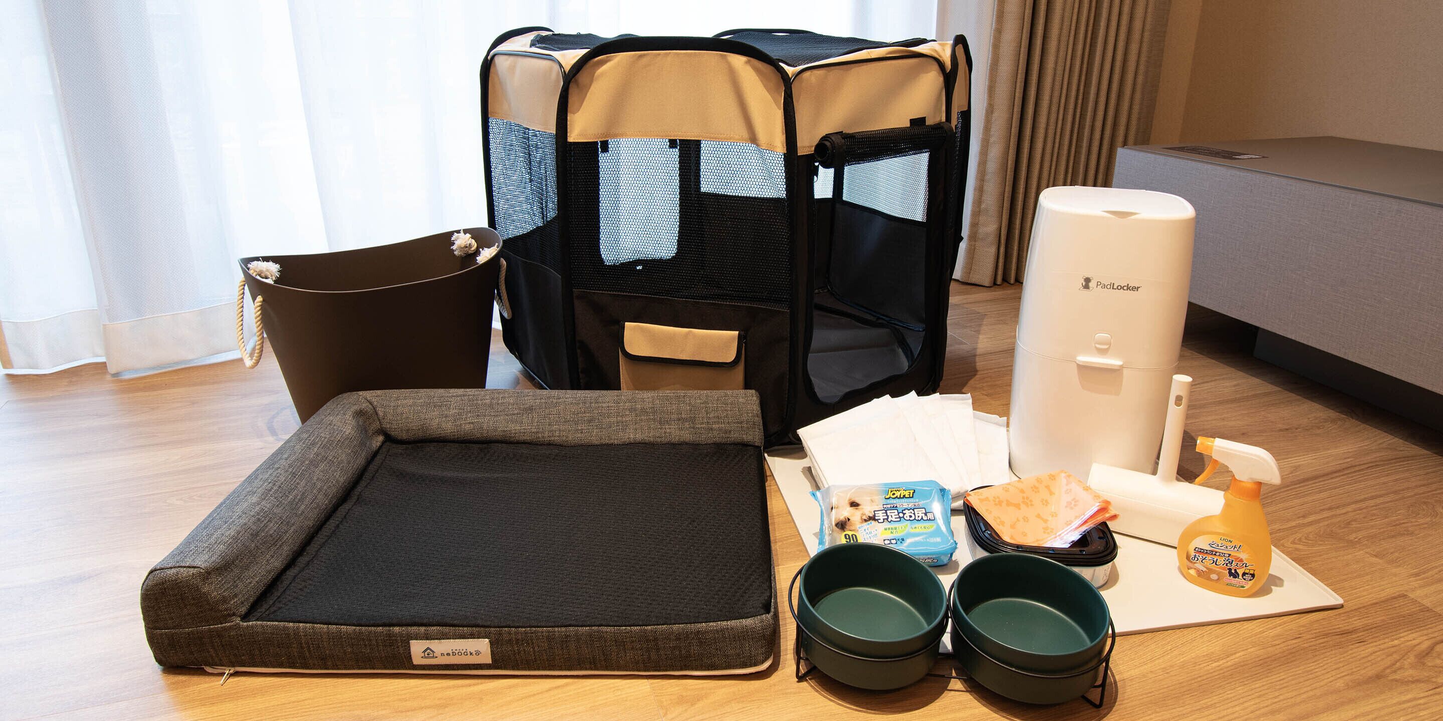 A pet bed, a pet carrier, bowls, a litter box, a litter box liner, a pet food package, and a spray bottle on a wooden floor.