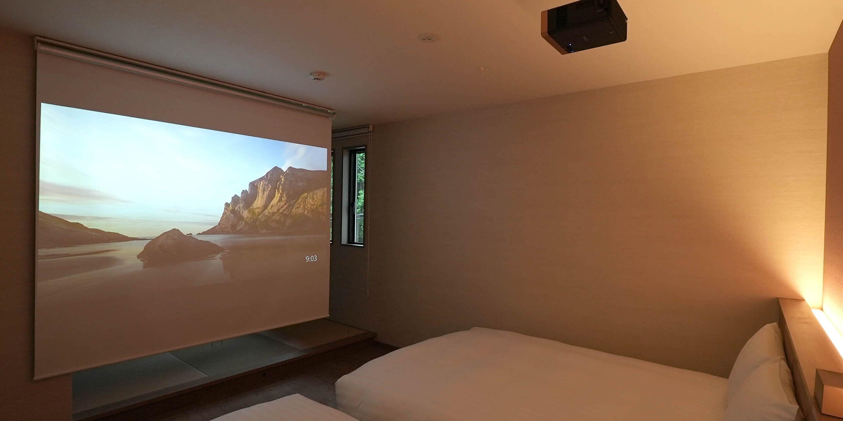 A hotel room with a bed, a projector screen displaying a scenic image, and a window with a view of the outdoors.